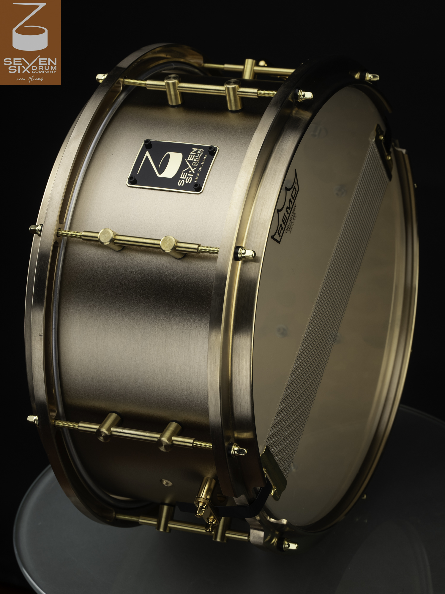 A snare drum featuring a brushed bronze shell with matching brass hoops, brass tube lugs, and bottom snare wires.