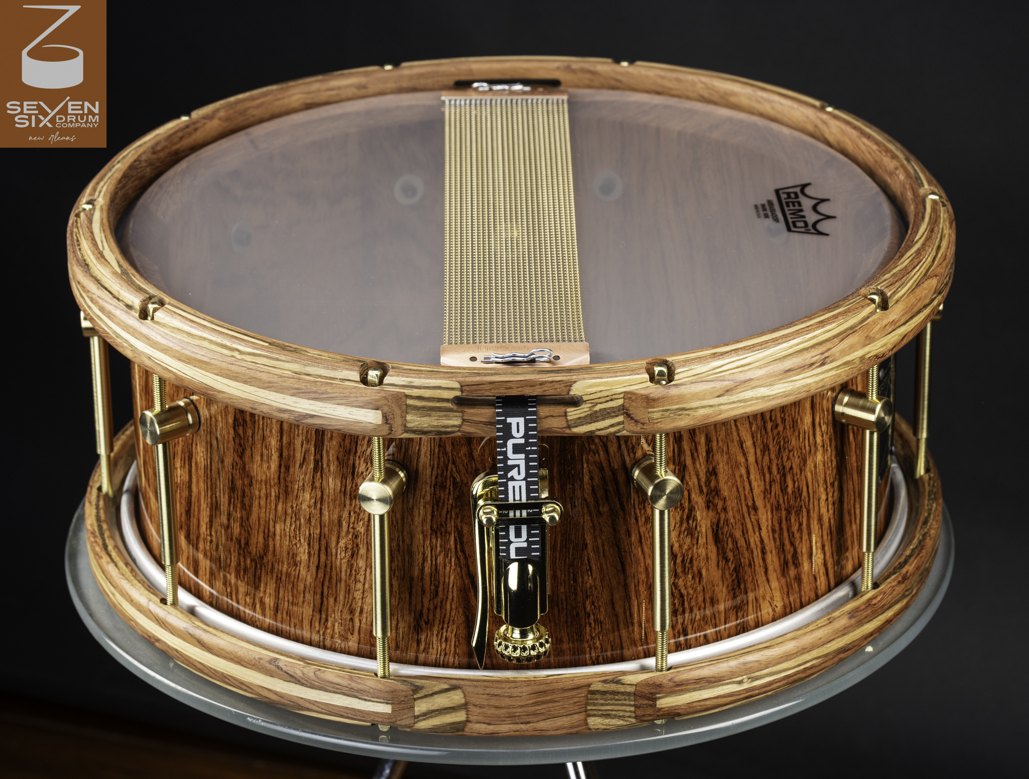 Handmade snare drum with a glossy exotic wood shell, segmented wooden hoops, brass tension rods, and snare wires.