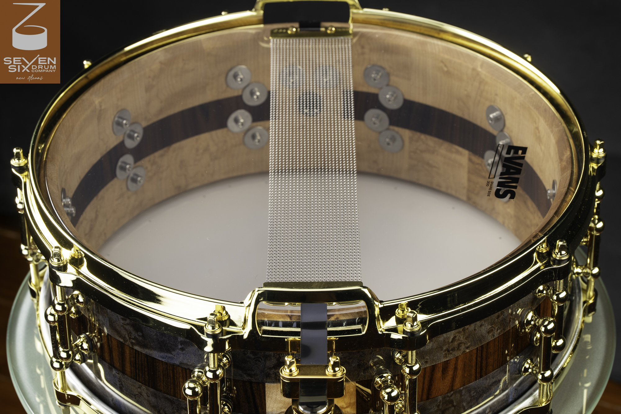 Handmade gloss wood snare drum featuring striped staves, polished gold hardware, and snare wires over a clear head.