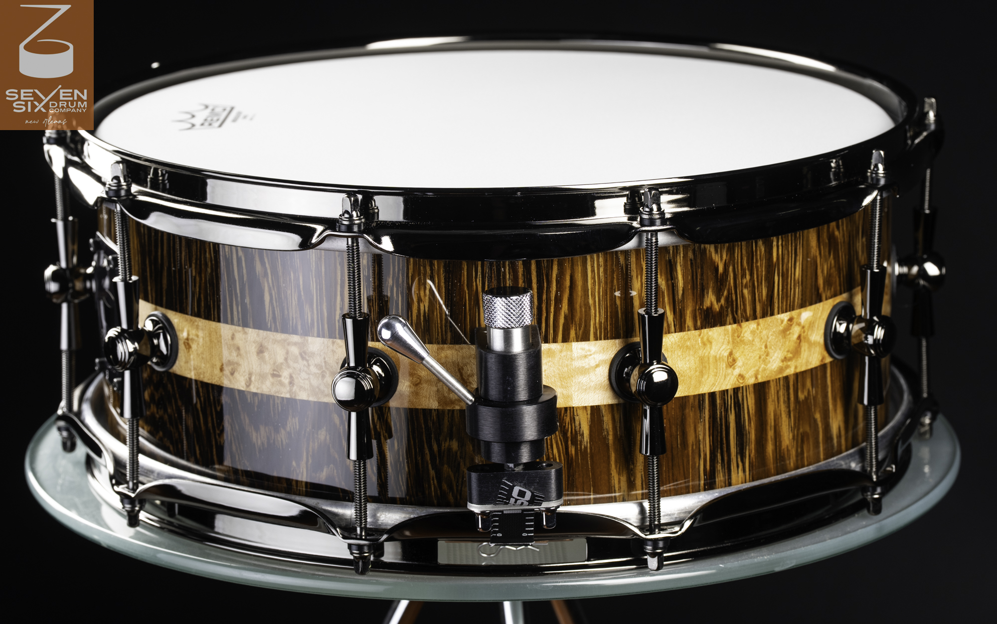 Handmade snare drum with a glossy wood grain shell, burl center inlay, black nickel hoops, and a throw-off strainer.