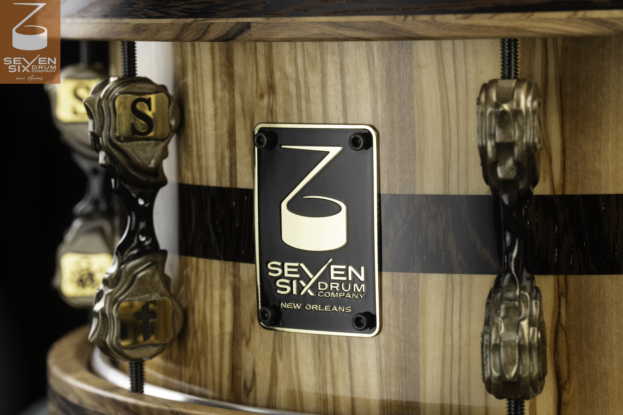 Natural wood snare drum with a dark stripe, gloss finish, custom antique brass lugs, and a black-and-gold maker's badge.