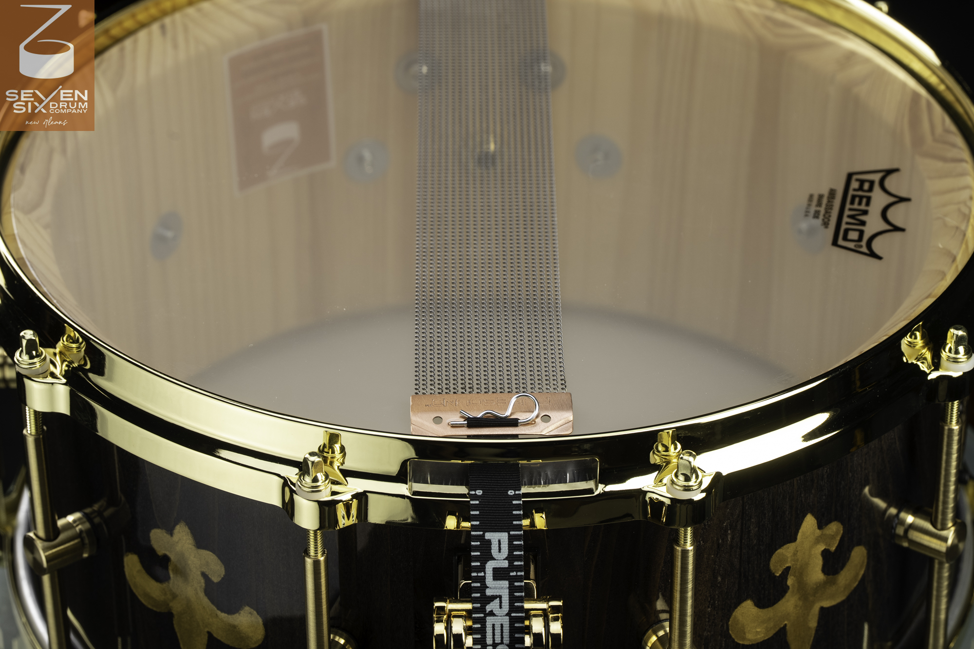 Underside of a dark wood snare drum with gold inlays, polished gold hardware, and wire snares across a clear head.
