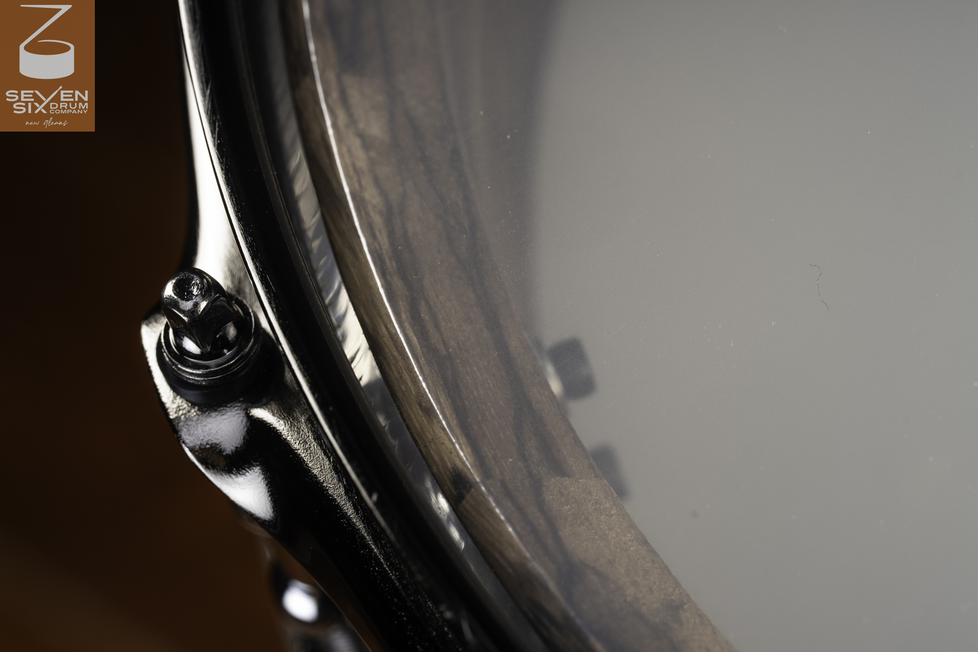 Close-up of a dark wood snare drum shell with a glossy black nickel hoop and tension rod under a clear drumhead.