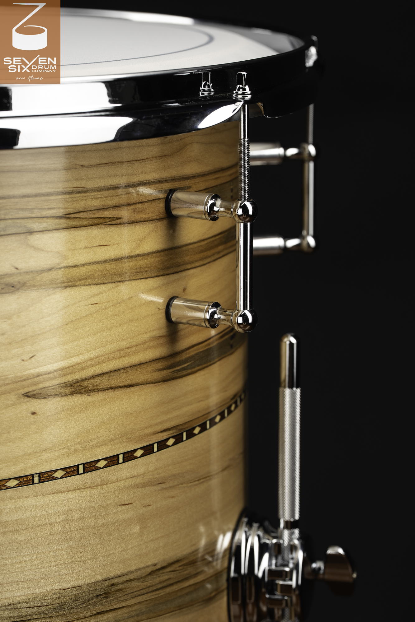Glossy natural wood snare drum with a geometric inlay band, chrome tube lugs, and a knurled throw-off lever.