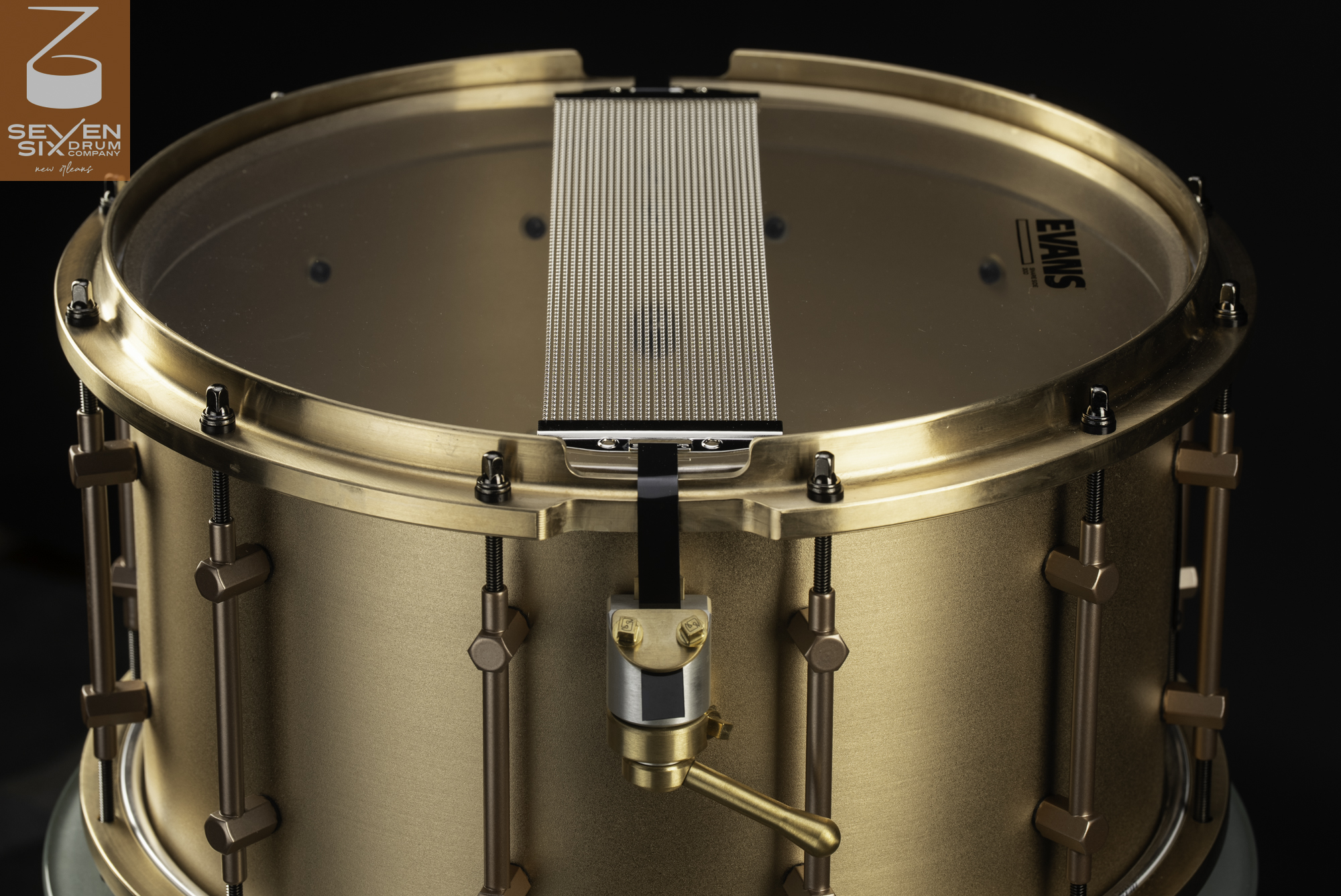 Brushed bronze snare drum bottom showing wire snares, matching tube lugs, and a snare throw-off mechanism.