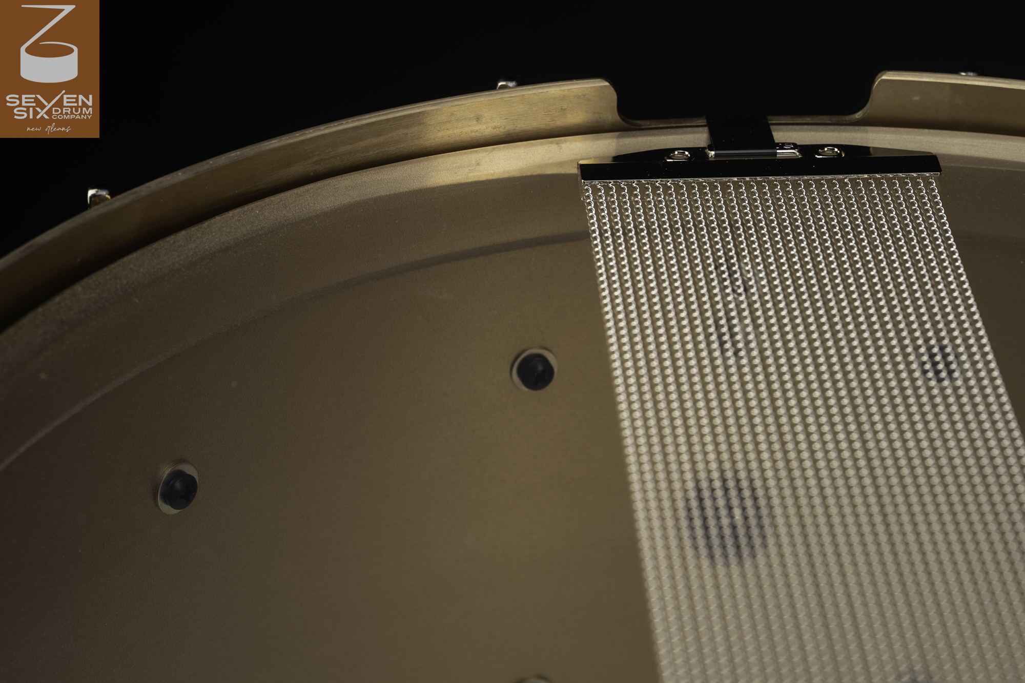 Bottom view of a brushed bronze snare drum with wide steel snare wires resting across a matching hoop.