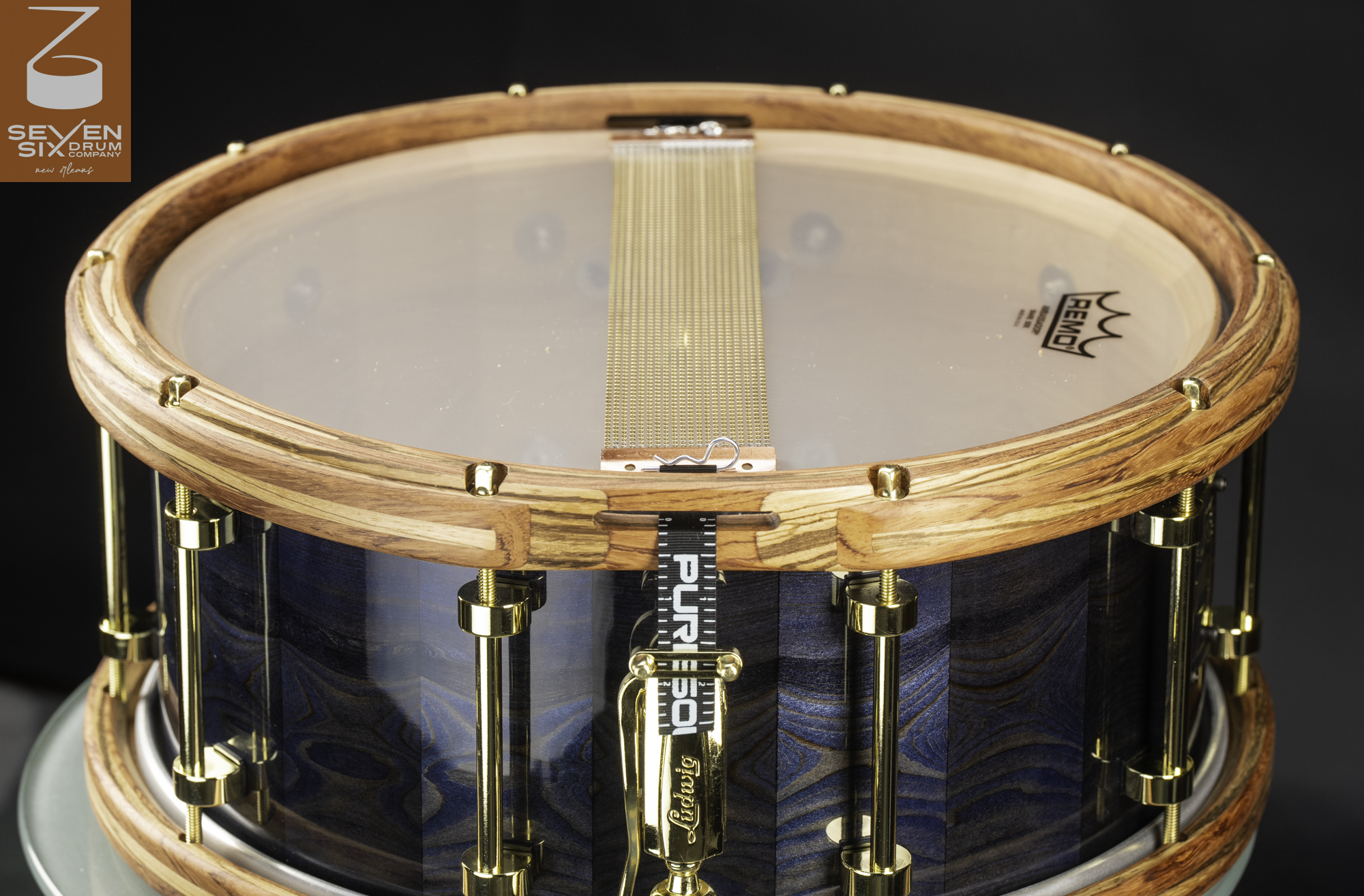 Handmade snare drum underside featuring a blue wood shell, natural wood hoops, gold tube lugs, and brass snare wires.