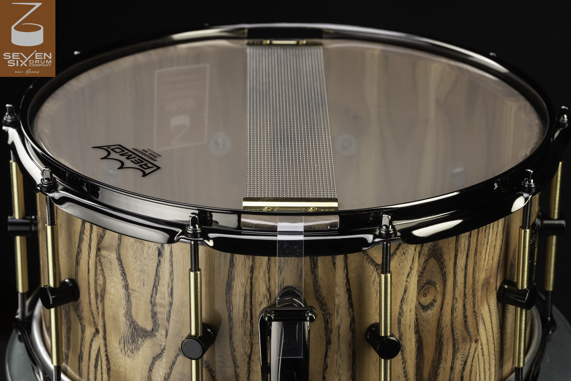 Natural wood snare drum with a clear finish, black hoops, brass tube lugs, and snare wires across a clear resonant head.