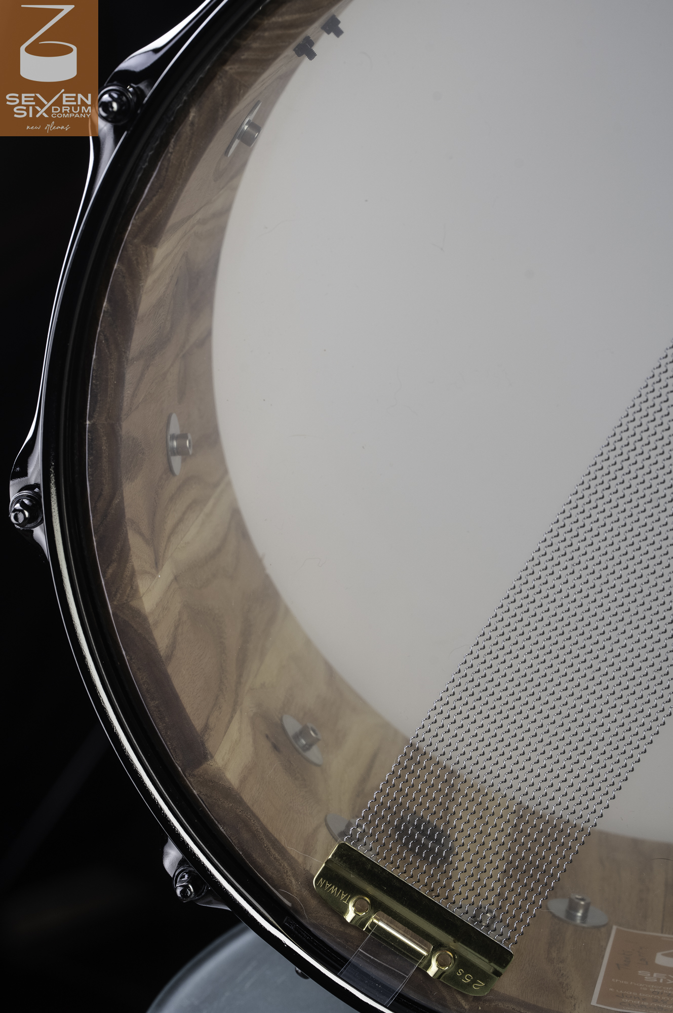 Interior of a natural-finish wood snare drum with a black hoop and steel snare wires secured with a brass clip.