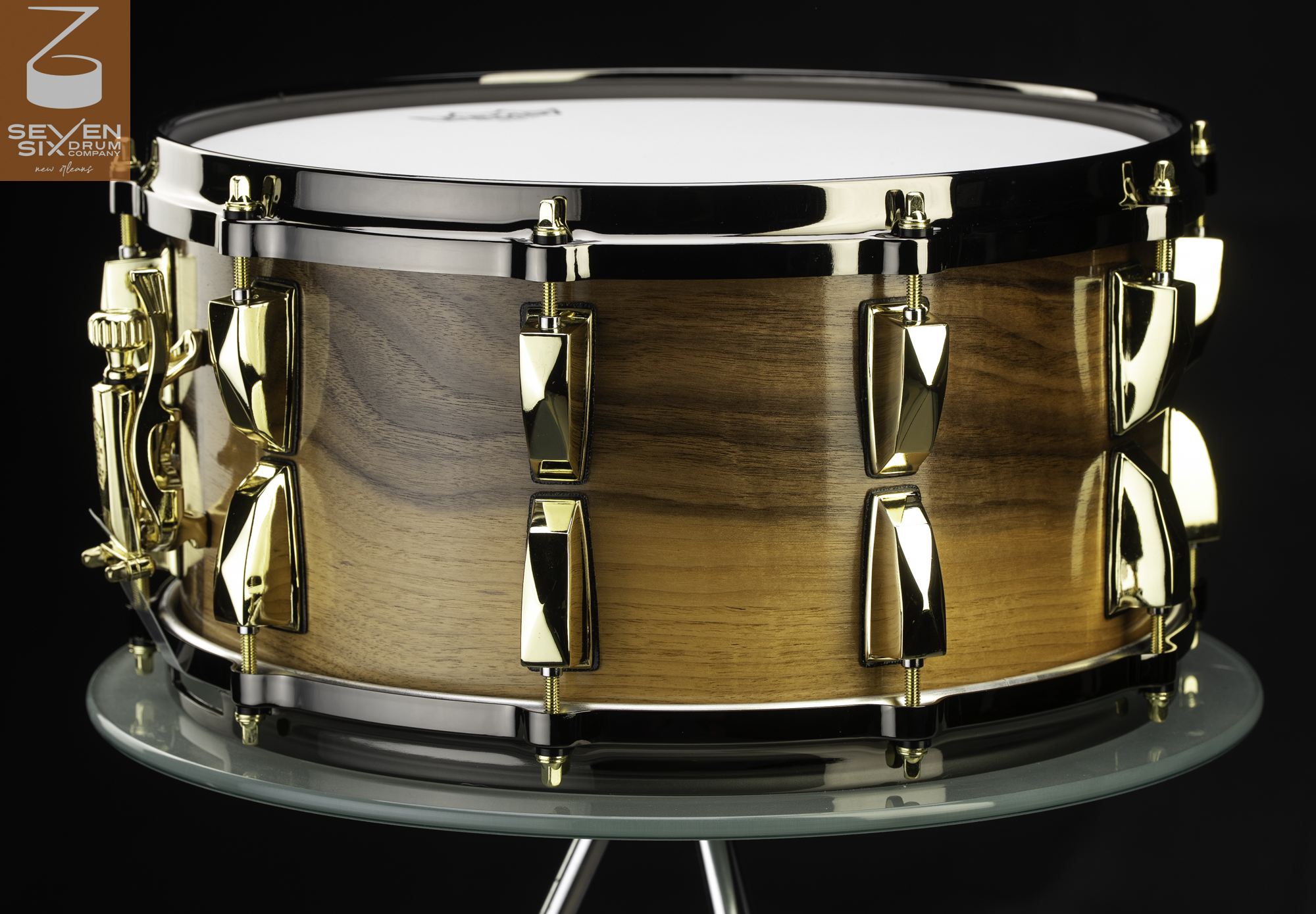 Handmade snare drum featuring a glossy wood shell, polished gold lugs, and black nickel hoops.