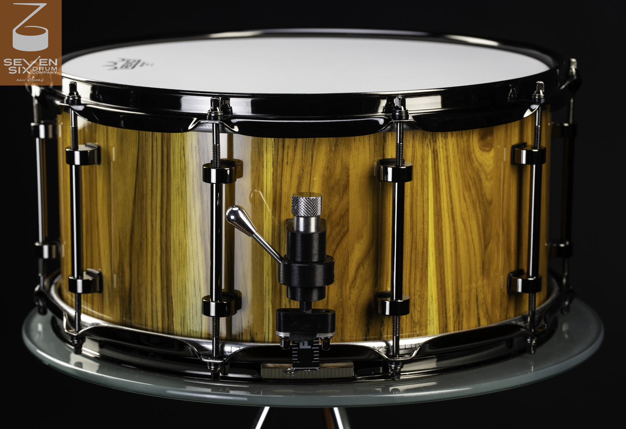 Handcrafted snare drum featuring a glossy golden wood shell, black nickel hardware, and an adjustable throw-off strainer.