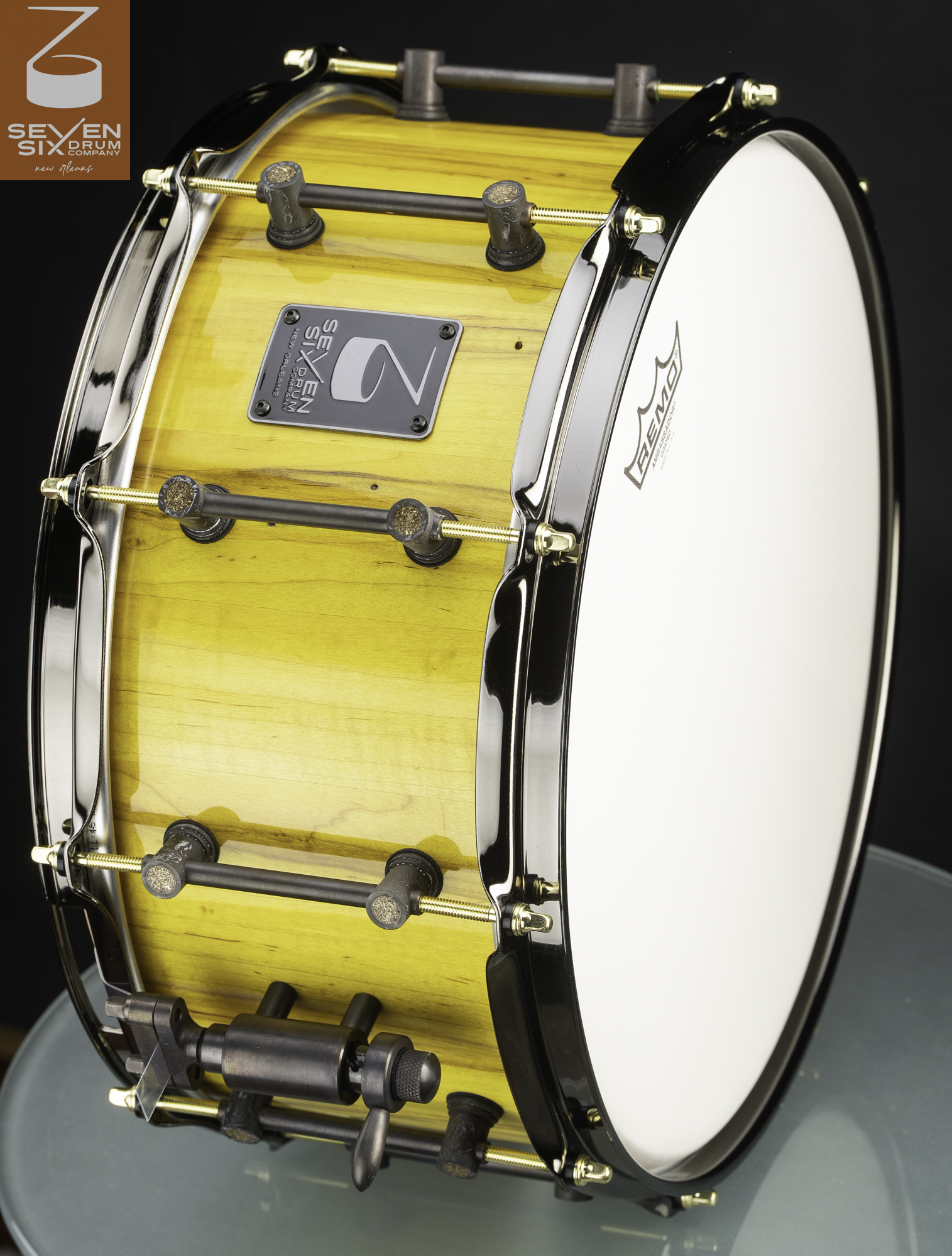 Glossy yellow wood snare drum with black chrome hoops, dark bronze tube lugs, and brass tension rods.