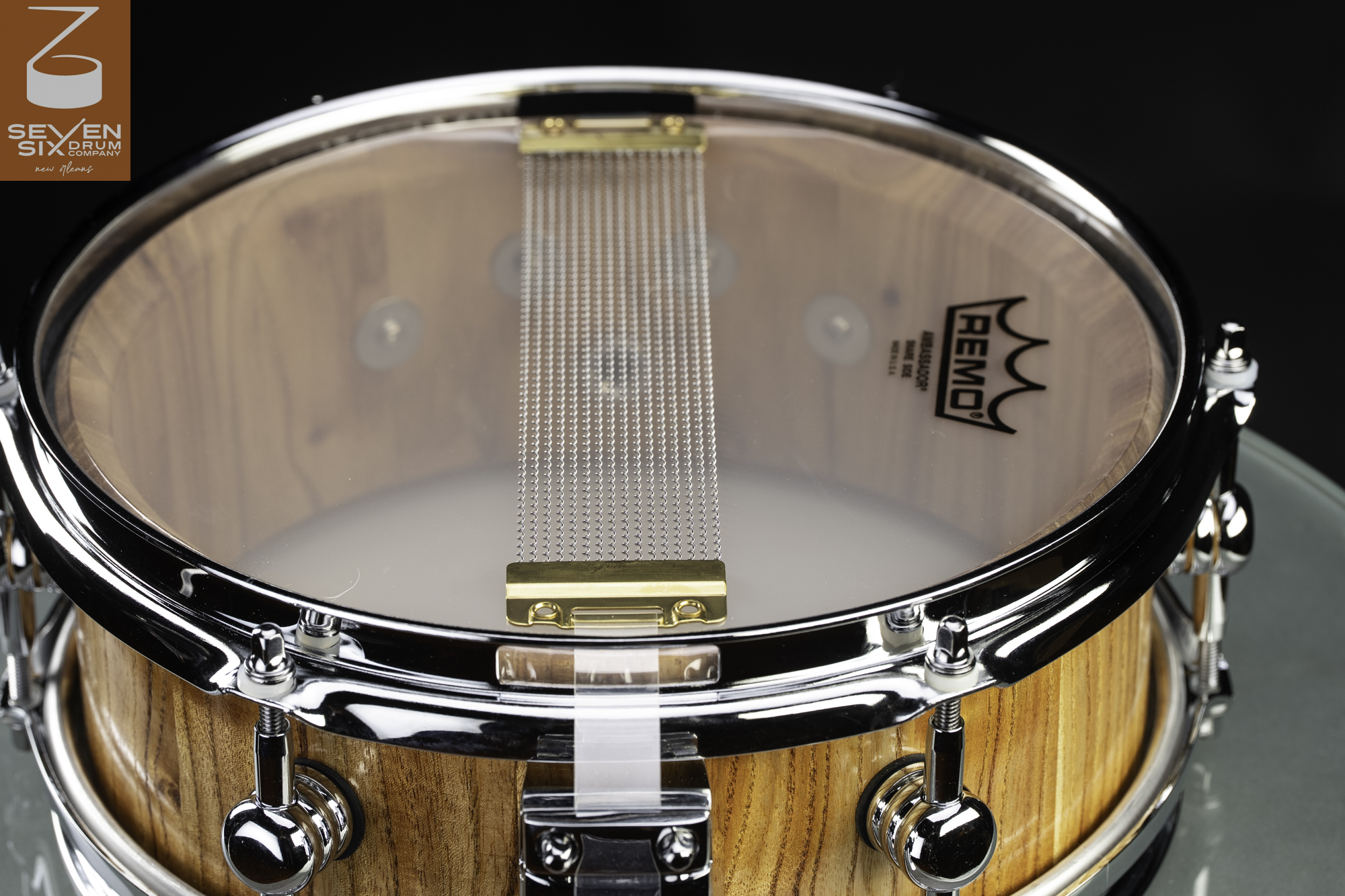 Underside of a natural wood snare drum featuring a clear drumhead, chrome hoops and lugs, and brass-ended snare wires.