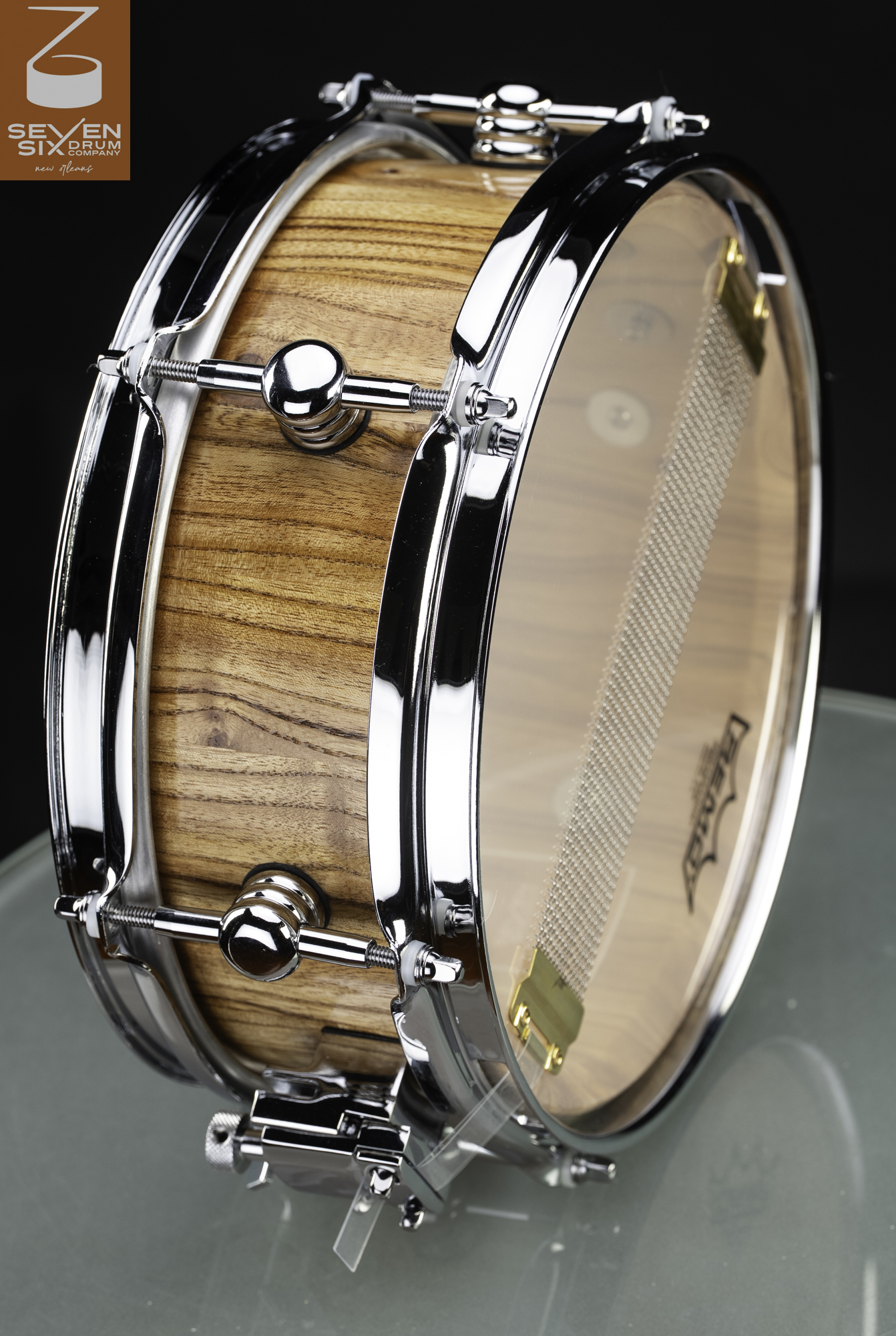 Handmade wood snare drum with a natural finish, chrome hoops, round lugs, and snare wires across a clear bottom head.