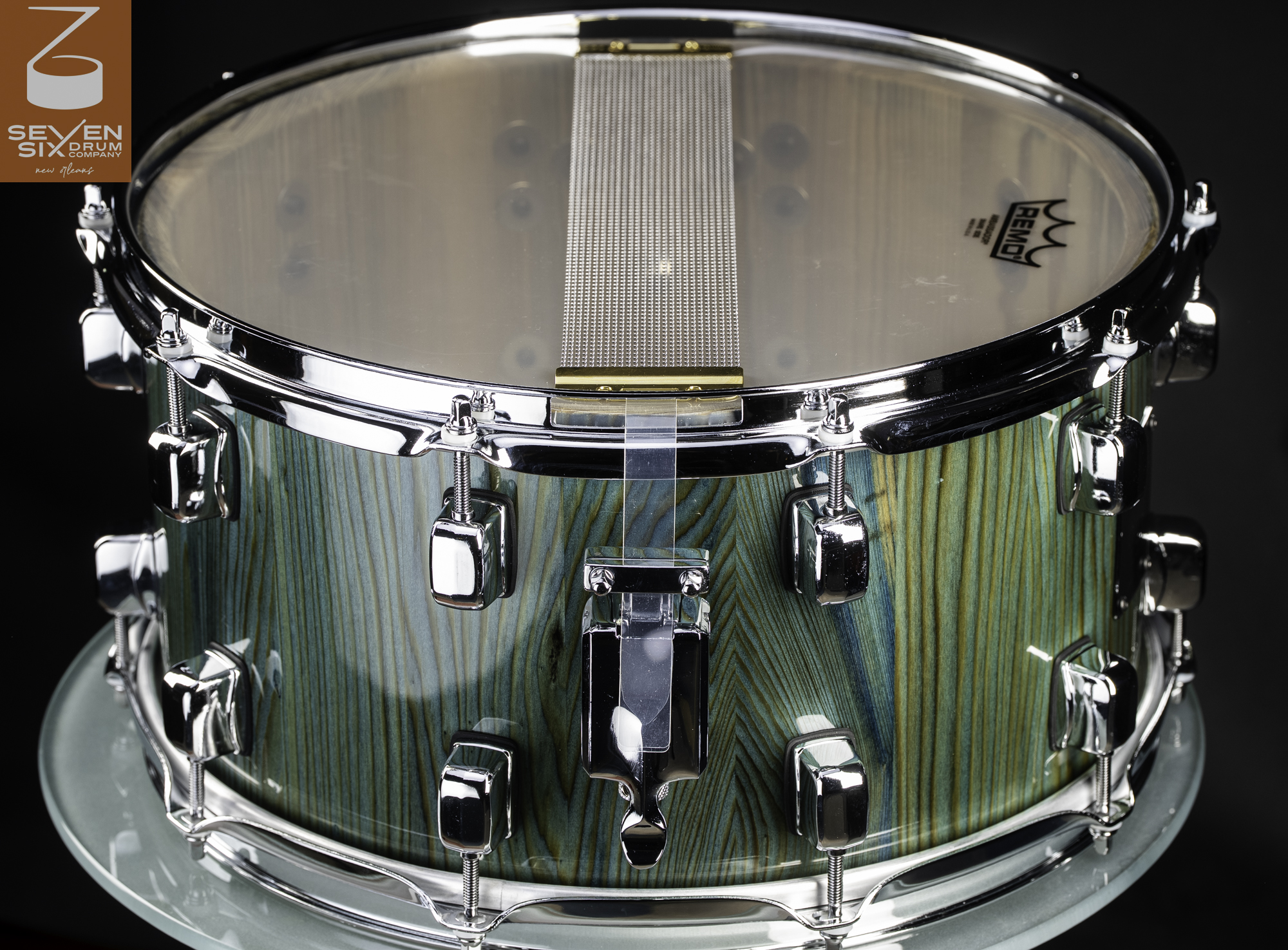 Handmade snare drum with a teal-stained wood finish, chrome lugs and hoops, and snare wires over a clear resonant head.
