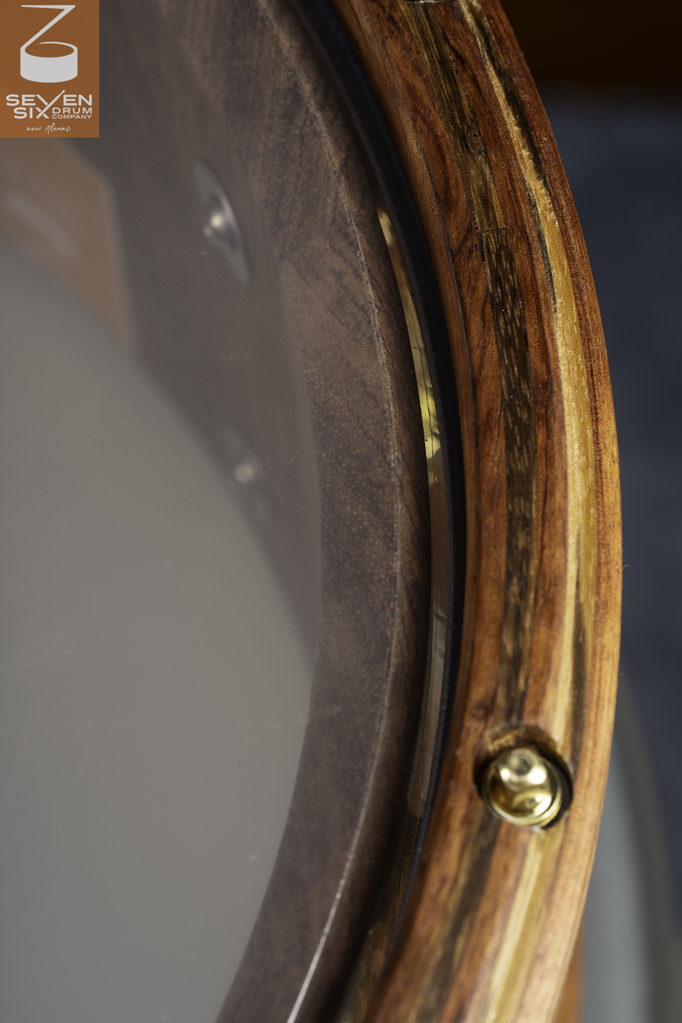 Close-up of a snare drum featuring a glossy layered wood hoop, dark wood shell, clear head, and recessed brass tension bolt.