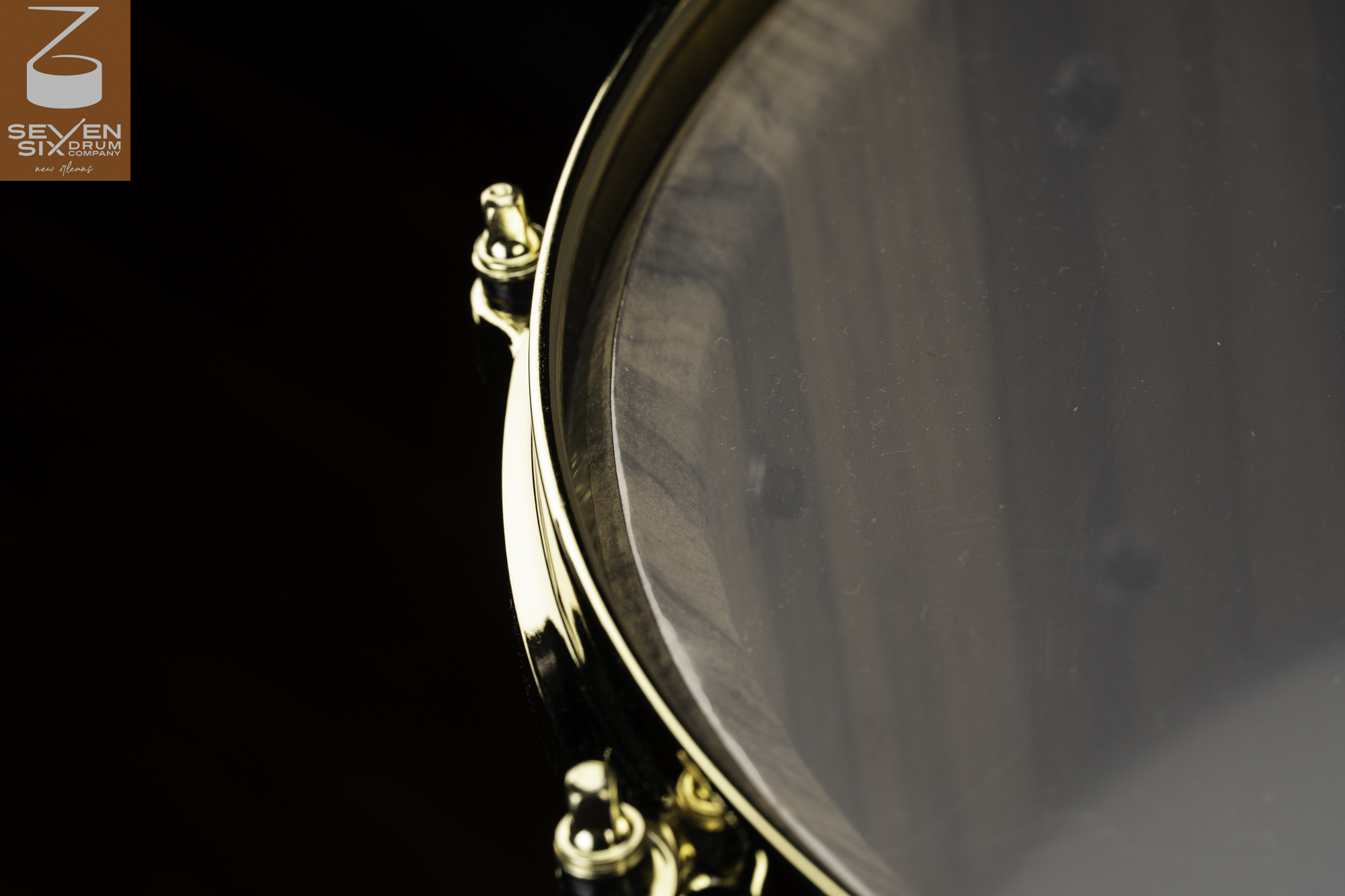 A handcrafted snare drum with a dark wood shell, clear head, and polished gold hoop and tension rods.