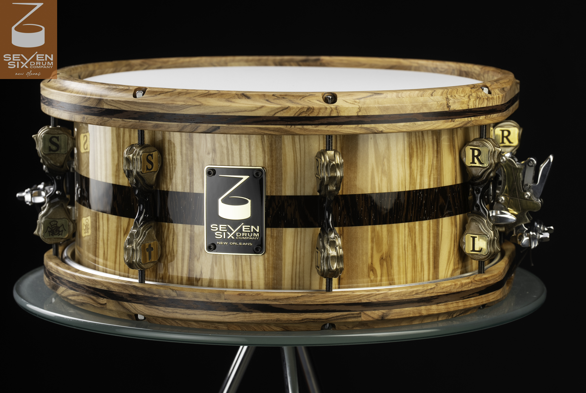 Handmade gloss Olivewood snare drum with a dark center Wenge stripe, matching Olive and Wenge wooden hoops, and sculpted bronze lettered lugs.