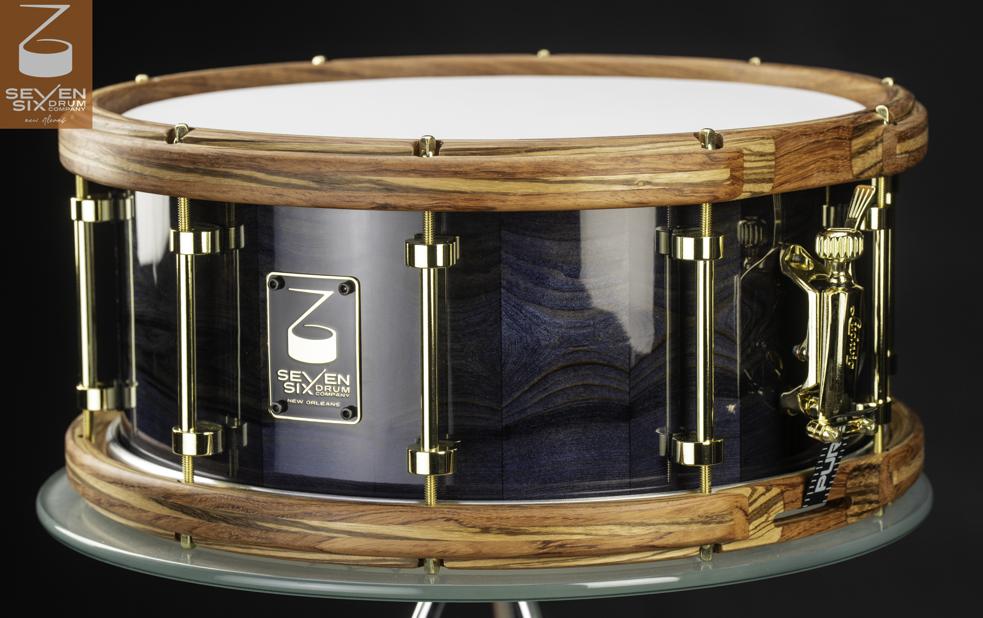 Handmade snare drum with a glossy blue Louisiana Cypress wood grain shell, nBubinga and zebrawood natural wood hoops, and polished gold tube lugs and strainer.