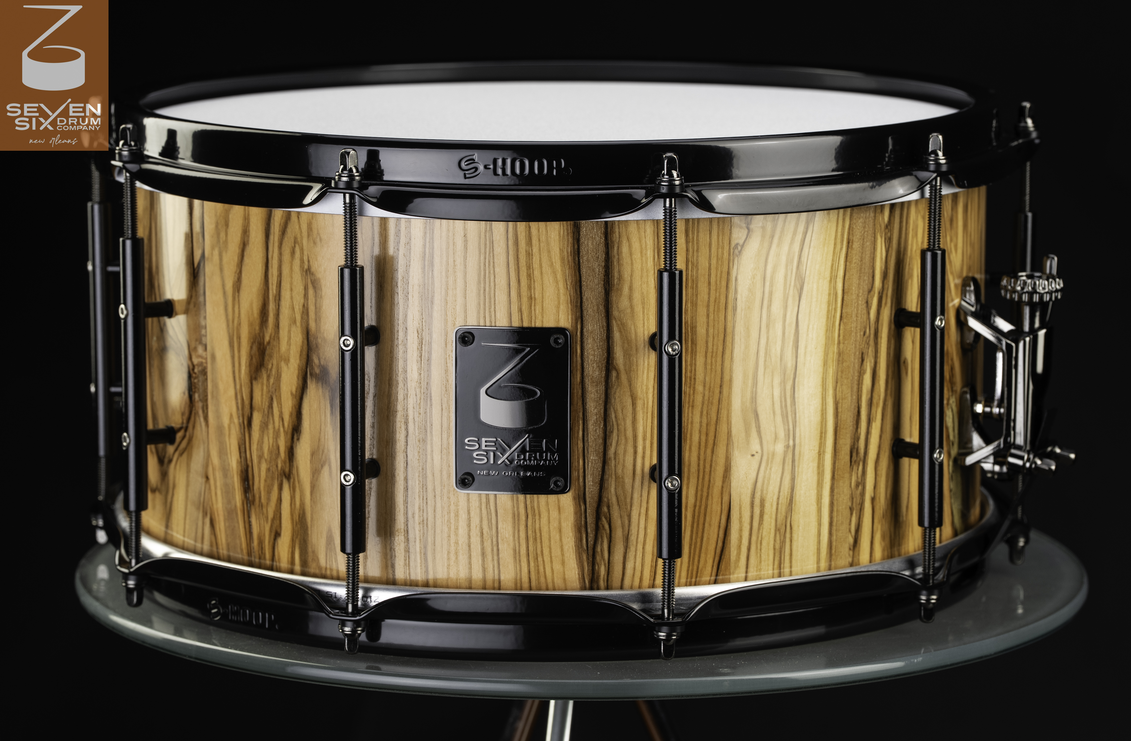 Handmade Olivewood snare drum featuring a glossy striped wood shell, black S-Hoop rims, and black tube lugs.